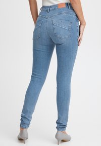 Mršave plave traperice s glatkom teksturom, visokim strukom, dva zadnja džepa i oznakom na pojasu. Dizajn koji prati nogu.