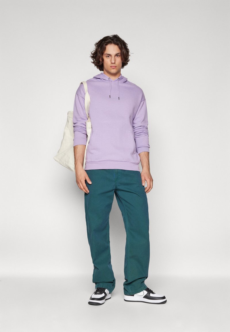 Felpa con cappuccio viola chiaro, pantaloni turchesi, sneakers bianco e nero, che tiene una borsa tote color crema, in piedi contro uno sfondo neutro.