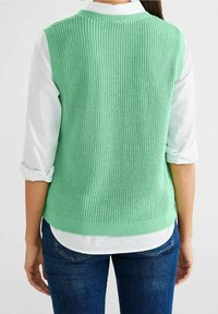 Mint green knitted sleeveless sweater with ribbed texture, worn over a white collared shirt, paired with blue jeans.