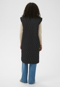 Long gilet noir matelassé avec un motif géométrique, manches courtes capuche, et une coupe droite, porté sur un haut beige et un jean bleu à jambes larges.
