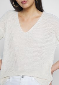 Biały sweter dzianinowy z dekoltem w serek i krótkimi rękawami, charakteryzujący się luźnym krojem i teksturowanym wzorem. Połączony z białymi spodniami w talii.