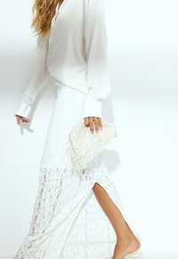 Blusa bianca trasparente con maniche lunghe, abbinata a una maxi gonna bianca di pizzo con uno spacco laterale. Tiene una borsa a tracolla bianca con frange.