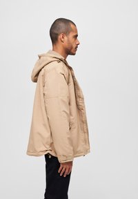 Beige hettejakke med glidelås foran, med en løs passform, lange ermer, sidelommer og en glatt, vannavstøtende tekstur.