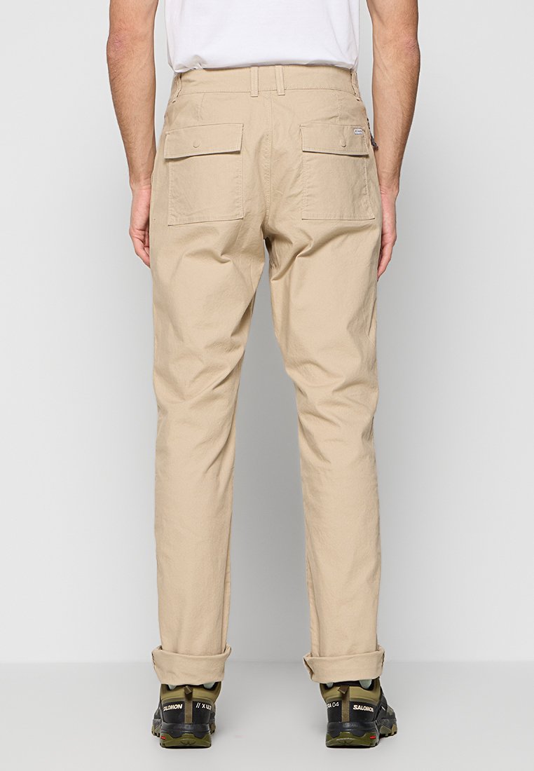 Pantalons cargo beiges avec deux poches arrière, ourlets retroussés et coupe décontractée, fabriqués en tissu léger. Associés à des chaussures de randonnée noires.