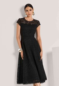 Schwarzes Spitzenkleid mit kurzen Ärmeln und tailliertem Oberteil, das mit floralen Ausschnitten und einem ausgestellten Rock versehen ist. Zubehör umfasst silberne Armbänder.