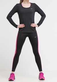 Mustapohjainen pitkähihaatinen urheilu-paita, jossa on pyöreä pääntie ja Nike-logo. Mustat legginsit, joissa on vaaleanpunaiset sivukohdat ja vaaleanpunaiset juoksukengät.