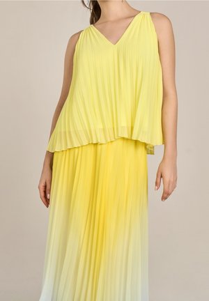 Vrouw die een mouwloze gele geplooide gelaagde jurk met V-hals draagt, staand tegen een neutrale achtergrond, armen ontspannen langs het lichaam.