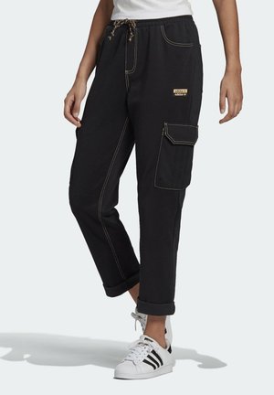 Personne portant un pantalon cargo noir avec des surpiqûres beiges et un petit logo Adidas sur la poche, assorti à des baskets blanches Adidas à rayures noires.