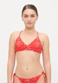 Rød bikini med hvitt blomstertrykk. Toppen har en trekantet fasong og tynne stropper; bunnen har snøring i sidene og et lignende blomsterdesign.