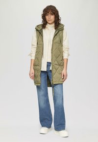 Quilted olivgrüne ärmellose Jacke mit Kapuze, kombiniert mit einem weiß gestreiften Hemd und blauen weiten Jeans, mit einem Reißverschluss.