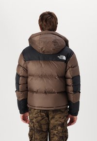 Brun och svart isolerad puffjacka med huva, med kviltade sektioner och en logotyp på övre ryggen. Bärs tillsammans med mönstrade cargobyxor.