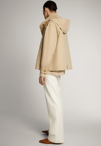 Beige hoodie met een ontspannen pasvorm, voorzien van knopenaccenten en een zachte textuur, gelaagd over een licht gebreid topje en gecombineerd met witte broeken.