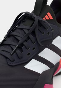 Chaussure de sport avec un dessus violet foncé en matériau texturé, accents blancs, lacets plats et contrefort de talon rose. Détails de logo visibles.