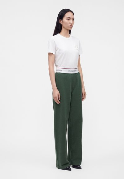 Femme aux longs cheveux noirs portant un t-shirt blanc ajusté et un pantalon vert taille haute à jambes larges avec un texte blanc sur la ceinture, debout immobile.