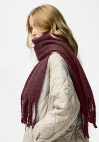 Burgunderfarvet uldslipset med frynser hængt over en lys quiltet jakke, hvilket fremhæver den bløde tekstur og den rige farvekraft.