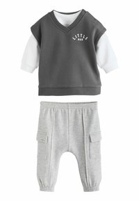 Grijze mouwloze top met de tekst "LITTLE MAN" boven een witte lange mouwen shirt, gecombineerd met lichtgrijze joggingbroek met zijzakken.