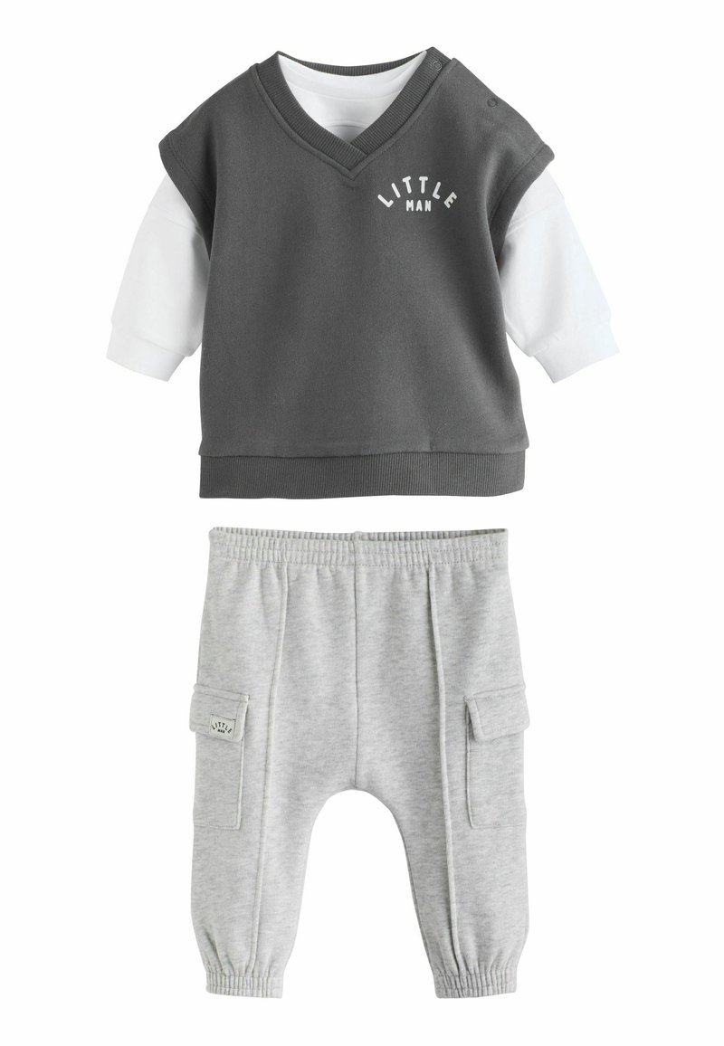 Grijze mouwloze top met de tekst "LITTLE MAN" boven een witte lange mouwen shirt, gecombineerd met lichtgrijze joggingbroek met zijzakken.