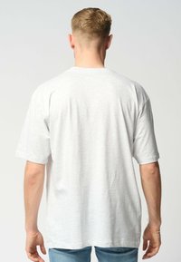 Mand med kort blondt hår iført en ensfarvet lys grå oversized T-shirt og blå jeans, stående med ryggen til mod en neutral baggrund.
