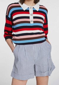 Gestreepte gebreide top in rood, blauw, roze en bruin met een witte kraag en decoratieve knopen; gecombineerd met blauw-witte gestreepte shorts met plooien.