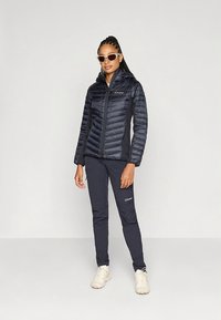 Teget jakna s prošivanim dijagonalnim uzorkom i patentnim zatvaračem. Uparena s teget hlačama i svijetlim cipelama. Dodaci uključuju sunčane naočale.