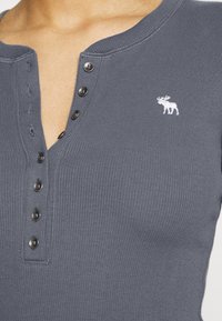 Grå ribbad henleytröja med V-ringning och fem svarta knappar. Har en liten vit broderad älglogo på vänster bröstområde.