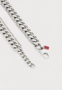 Hõbedast kettkaelakee, mis koosneb laiematest, tekstuuriga metallist segmentidest, koos väikese punase sildi lisandiga sulgemise lähedal.