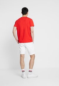 Červené bavlněné tričko, krátké rukávy, pohled zezadu. Nositeli s bílými šortkami, bílými teniskami a pruhovanými ponožkami. Hladká textura, bez viditelných log.