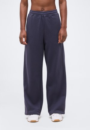 Άτομο που φοράει φαρδιά σκούρα μπλε παντελόνια και λευκά αθλητικά παπούτσια, στέκεται μπροστά σε ανοιχτό γκρι φόντο με εστίαση στον κάτω κορμό και τα πόδια.