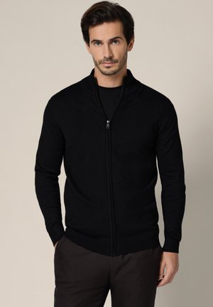 Mężczyzna o krótkich ciemnych włosach, ubrany w czarny sweter na zamek i ciemne spodnie, stojący na tle jednolitego jasnego tła.