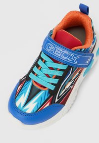 Pantof sportiv albastru și negru cu partea superioară modelată cu desene zigzag, șireturi turcoaz, accente roșii și o curea cu velcro.