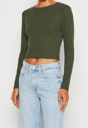 Vrouw draagt een aangesloten olijfgroen cropped top met lange mouwen, gecombineerd met lichtblauwe, hooggeleide jeans.
