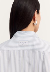 A person with long dark hair wears a light blue striped shirt, showcasing a label that reads "per sempre tita" and a large silver earring.