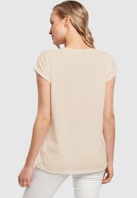 Beige t-skjorte med korte ermer og rulleermer, myk stoff, avslappet passform og rund hals, kombinert med hvite bukser.
