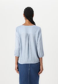 Blusa blu chiaro con maniche 3/4, caratterizzata da un motivo a quadri e un colletto rotondo. Il retro presenta un delicato dettaglio a piega.