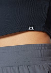 Crna rebrasta kratka majica prelazi u sive hlače s teksturiranim pojasom. Crna oznaka Under Armour vidljiva je na rubu majice.
