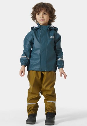 Chaqueta impermeable color teal con capucha, detalles reflectantes, combinada con pantalones impermeables marrones y botas negras. El niño está de pie con las manos relajadas.