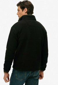 Sort fleece jakke med høj krave, tekstureret overflade og rektangulær rygpanel. Båret med blå jeans, hvor ærmerne er delvist oprevet.