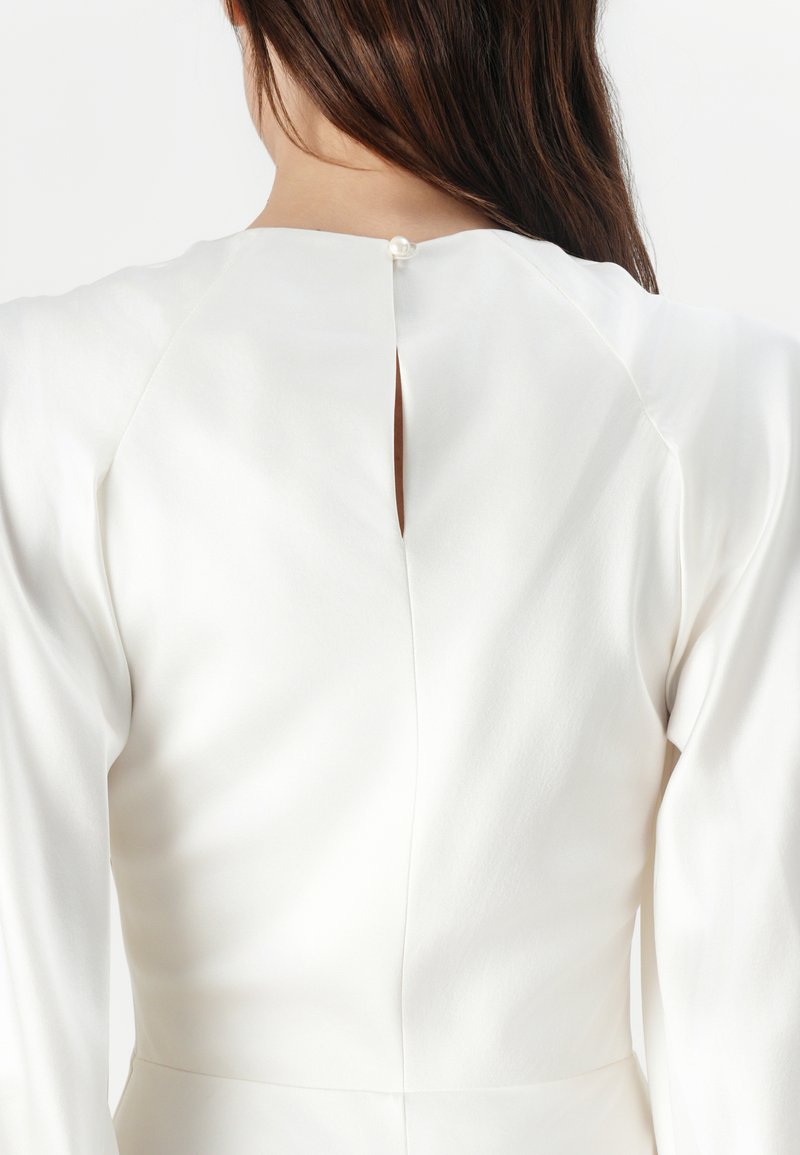 Robe blanche en tissu lisse, avec des manches longues, une petite ouverture en forme de clef dans le dos, et un bouton décoratif en perle au niveau du col.