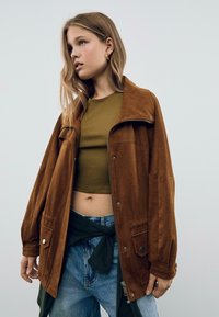 Veste en daim marron au style décontracté, dotée d'un col, d'une fermeture zippée et de grandes poches. Associée à un crop top vert olive et à un jean bleu.