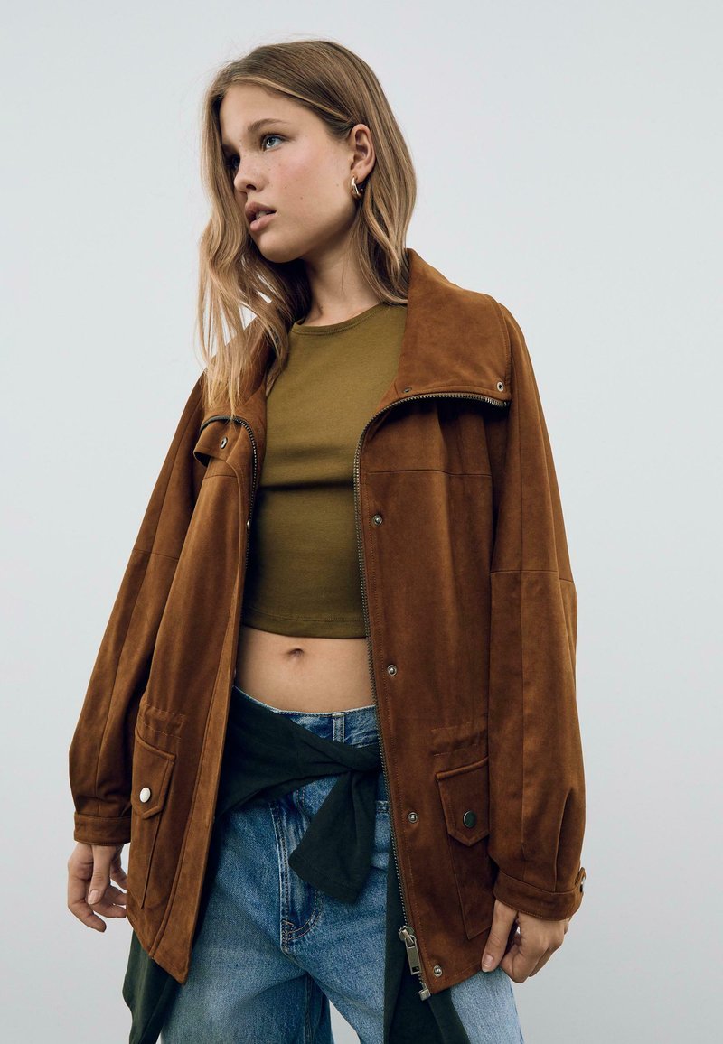 Veste en daim marron au style décontracté, dotée d'un col, d'une fermeture zippée et de grandes poches. Associée à un crop top vert olive et à un jean bleu.