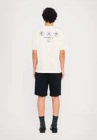 Cream short-sleeve t-shirt with back print featuring “EA7” and logo. Paired with black shorts and black shoes, model stands facing away.