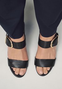 Schwarze Ledersandalen mit einem breiten Zehenriemen, einem Knöchelriemen mit goldener Schnalle und flacher Sohle. Glatte Textur und minimalistisches Design.