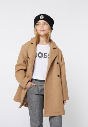 Tan wolmix jas met een dubbelgebreide ontwerp, zwarte knopen, gedragen over een witte cropped t-shirt met zwarte grafische tekst, gecombineerd met grijs patroonbroek.