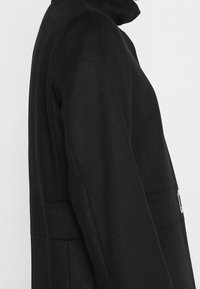 Cappotto in misto lana nera con colletto alto, texture liscia e dettaglio della cintura laterale. Presenta una silhouette strutturata e un design minimalista.