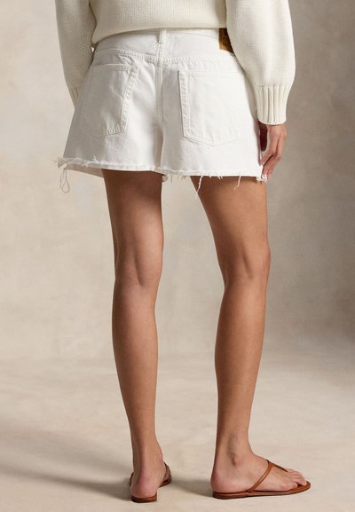 Γυναίκα με λευκά ξεφτισμένα τζιν σορτς, εκρού πουλόβερ και καφέ επίπεδα σανδάλια, εμφανιζόμενη από πίσω σε ουδέτερο φόντο.