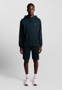 Felpa verde scuro con tasca a marsupio frontale e logo giallo, abbinata a pantaloni corti coordinati. Il modello indossa scarpe da ginnastica bianche e calzini.