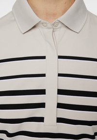Jasno beżowa koszulka polo z dwiema guzikami, w poziome paski na przemian czarne i białe. Miękki, teksturowany materiał.