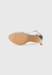 Silberne High-Heels mit glänzender metallischer Oberfläche, Riemen und einer strukturierten Gummisohle. Verfügt über eine abgerundete Zehenpartie und einen schlanken Absatz.