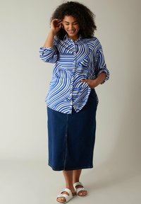 Camicia a righe blu e bianche con vestibilità comoda e maniche arrotolate, abbinata a una gonna di denim scuro e sandali bianchi.