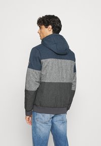 Color-blocked hooded jacket in navy, gray, and black hues; textured fabric with a ribbed hem; contrasting sleeves and a drawstring hood.